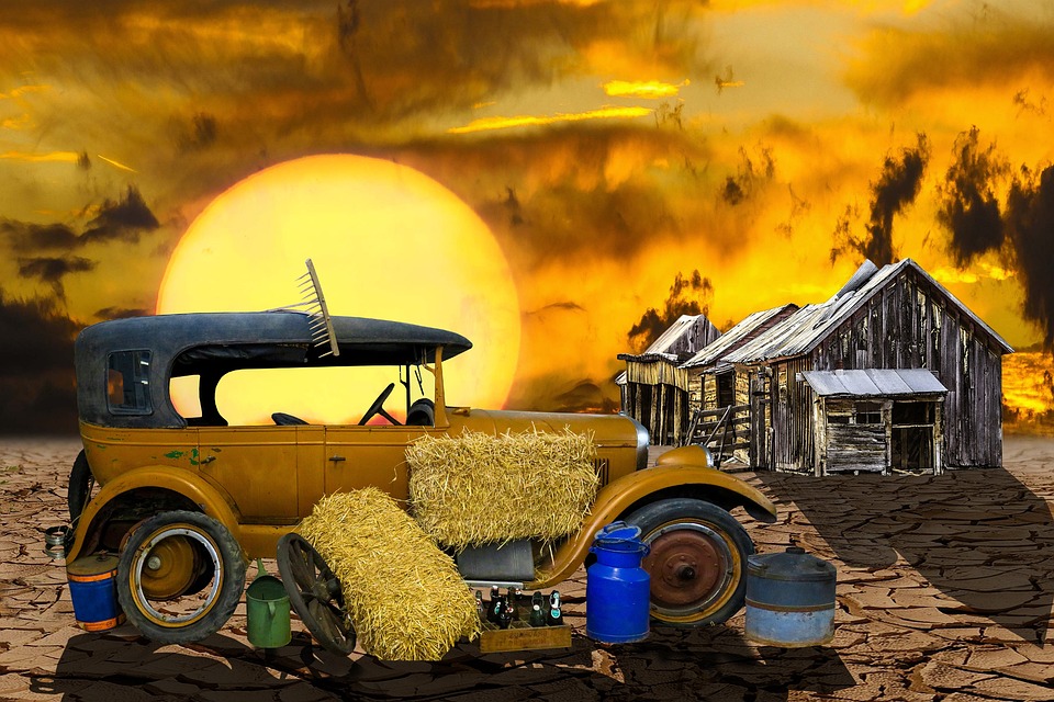Vintage car with hay bales parked on cracked ground near dilapidated wooden sheds under a dramatic sunset sky, illustrating the impact of climate.