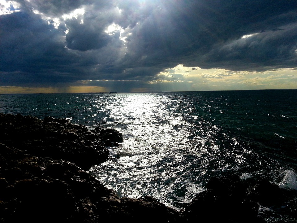 Sunlight piercing through dark clouds over a rocky shoreline with a shimmering sea, reflecting the changing climate.