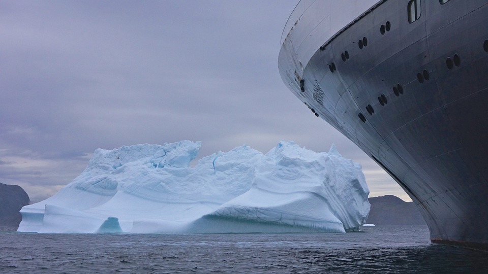 Cruise ship bow approaching a large iceberg in a climate-affected misty sea environment.