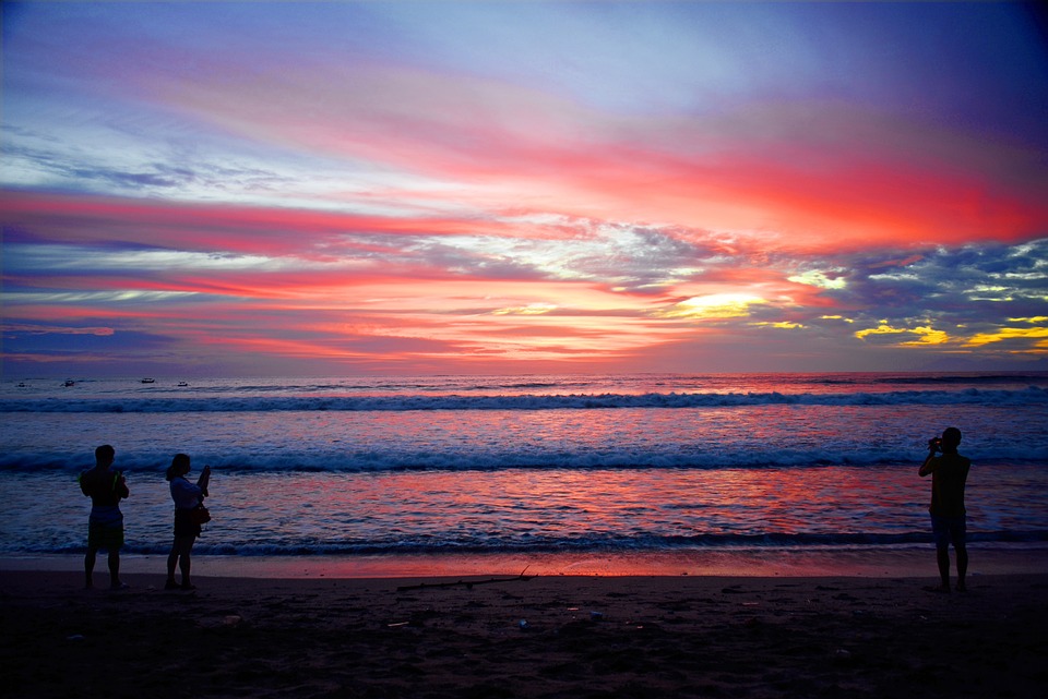 Three individuals on a beach capturing the vibrant sunset with their phones, documenting the climate's influence on the day's end.