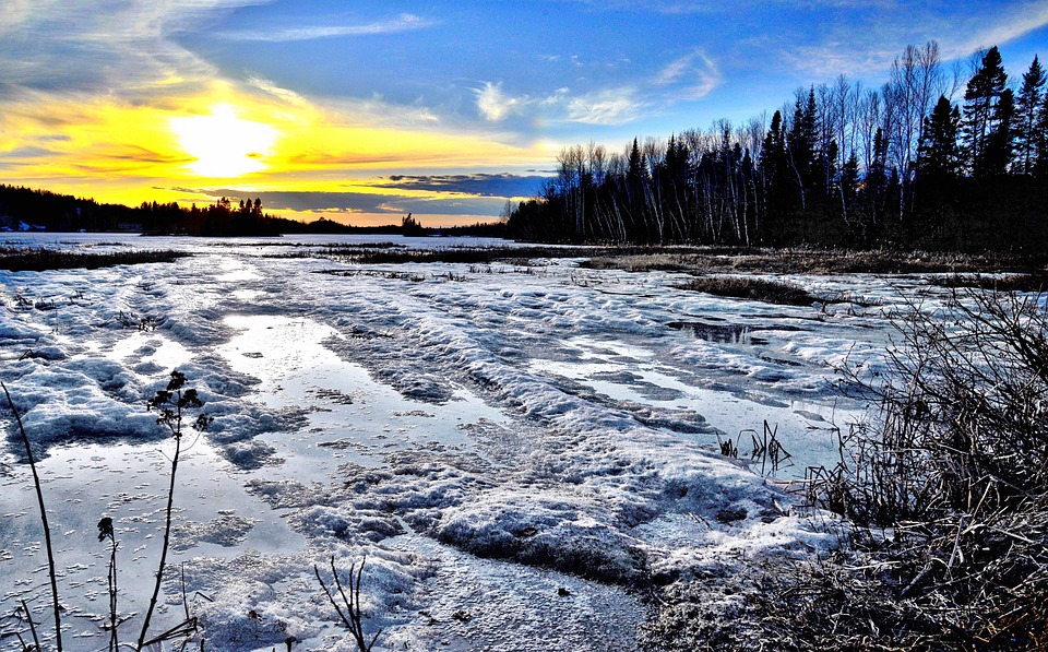 A winter sunset over a snow-covered landscape with icy paths and bare trees, depicting the cold climate.