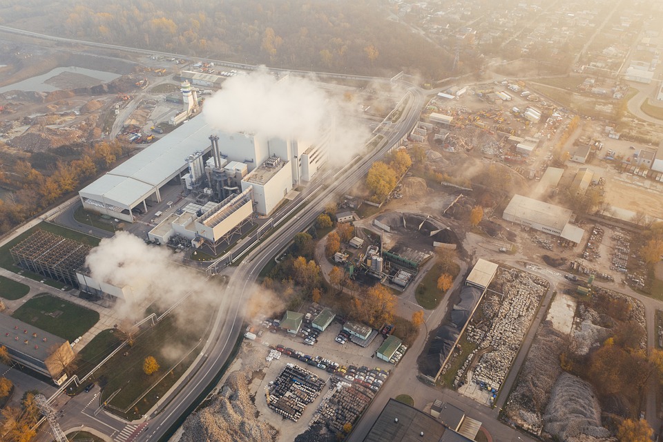 Aerial view of an industrial facility emitting steam or smoke, contributing to climate issues, surrounded by roads and smaller buildings.